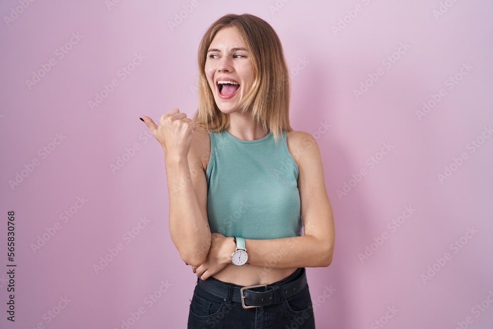 Blonde caucasian woman standing over pink background smiling with happy face looking and pointing to the side with thumb up. Blonde caucasian woman standing over pink background smiling with happy face looking and pointing to the side with thumb up.