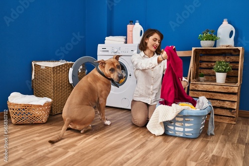 Young caucasian woman washing clothes sitting on floor with dog at laundry room
