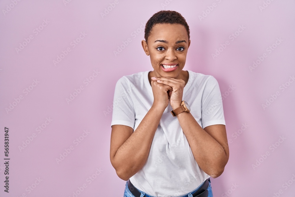 Beautiful african american woman standing over pink background laughing nervous and excited with hands on chin looking to the side Beautiful african american woman standing over pink background laughing nervous and excited with hands on chin looking to the side