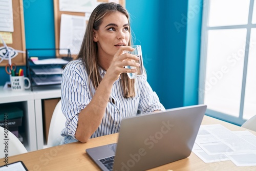 Young beautiful hispanic woman business worker using laptop drinking water at office
