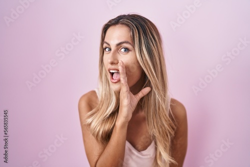 Young blonde woman standing over pink background hand on mouth telling secret rumor, whispering malicious talk conversation