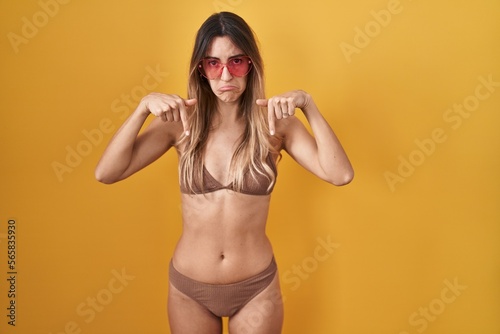 Young hispanic woman wearing bikini over yellow background pointing down looking sad and upset, indicating direction with fingers, unhappy and depressed.