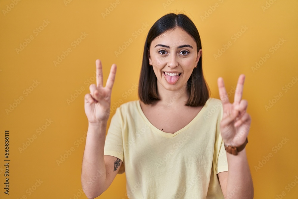 Hispanic girl wearing casual t shirt over yellow background smiling with tongue out showing fingers of both hands doing victory sign. number two. Hispanic girl wearing casual t shirt over yellow background smiling with tongue out showing fingers of both hands doing victory sign. number two.