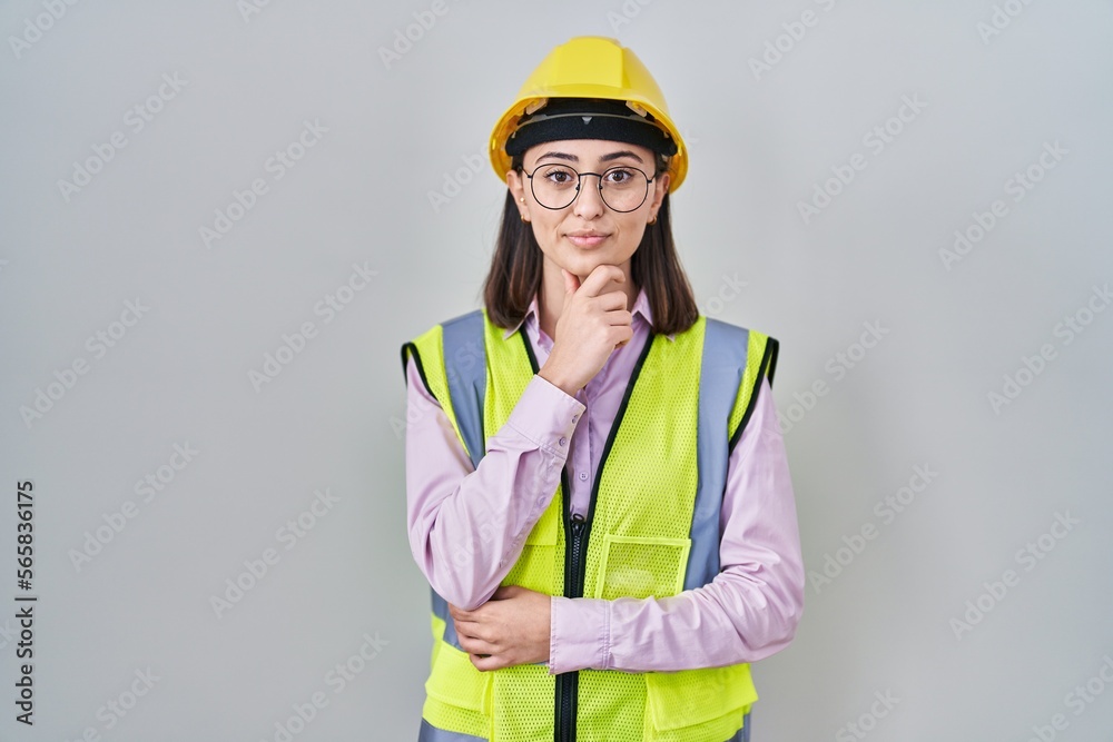 Hispanic girl wearing builder uniform and hardhat looking confident at the camera with smile with crossed arms and hand raised on chin. thinking positive. Hispanic girl wearing builder uniform and hardhat looking confident at the camera with smile with crossed arms and hand raised on chin. thinking positive.