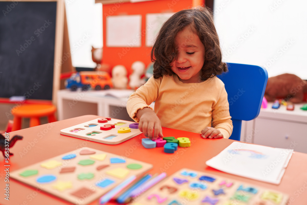 Adorable hispanic toddler playing with maths puzzle game sitting on table at kindergarten