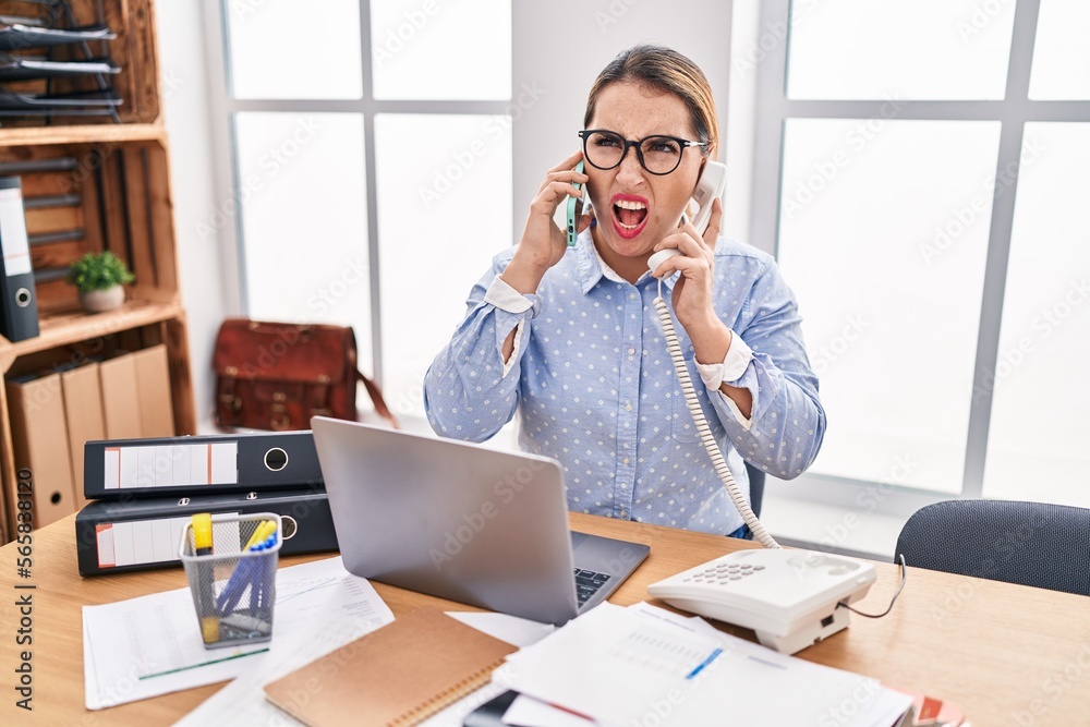Young hispanic business woman working at the office talking on two phones angry and mad screaming frustrated and furious, shouting with anger. rage and aggressive concept. Young hispanic business woman working at the office talking on two phones angry and mad screaming frustrated and furious, shouting with anger. rage and aggressive concept.