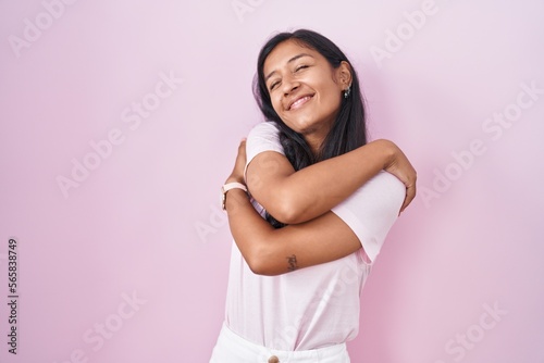 Fotografia Young hispanic woman standing over pink background hugging oneself happy and positive, smiling confident