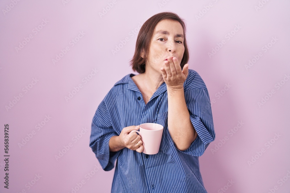 Middle age hispanic woman drinking a cup coffee looking at the camera blowing a kiss with hand on air being lovely and sexy. love expression. Middle age hispanic woman drinking a cup coffee looking at the camera blowing a kiss with hand on air being lovely and sexy. love expression.