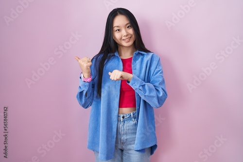 Young asian woman standing over pink background pointing to the back behind with hand and thumbs up, smiling confident