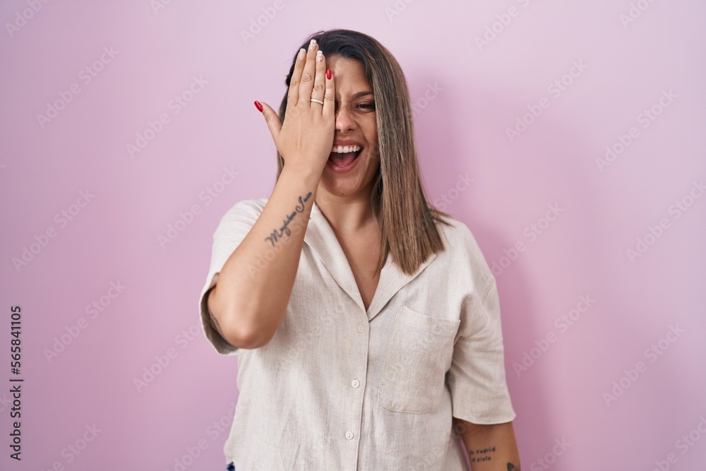 Blonde woman standing over pink background covering one eye with hand, confident smile on face and surprise emotion. Blonde woman standing over pink background covering one eye with hand, confident smile on face and surprise emotion.