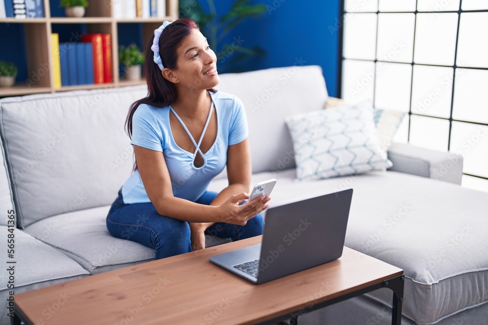 Obraz premium Young beautiful hispanic woman using smartphone and laptop at home