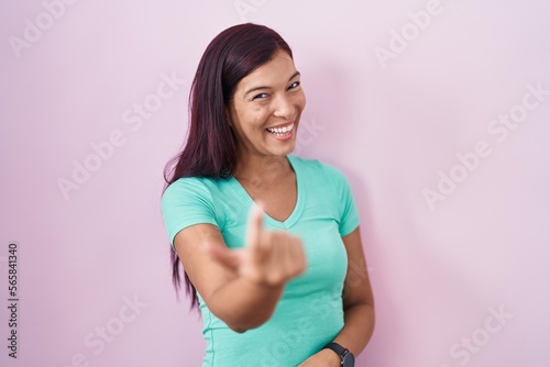 Young hispanic woman standing over pink background beckoning come here gesture with hand inviting welcoming happy and smiling