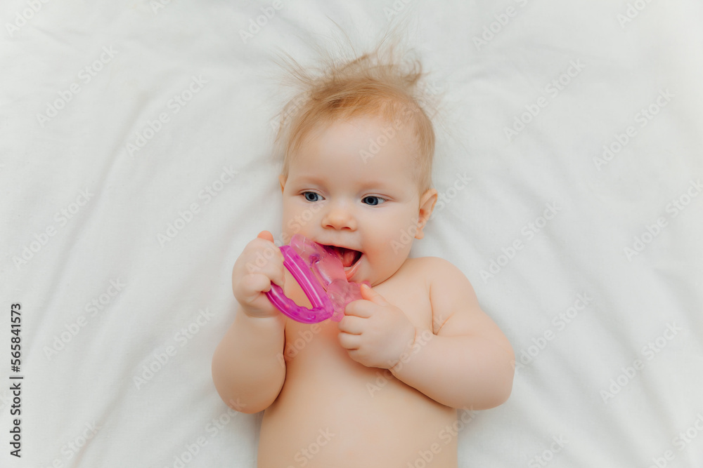 The child grows the first tooth. Smiling baby 6 months old chewing teether in bed closeup