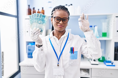 African woman with braids working at scientist laboratory holding money smiling looking to the side and staring away thinking.