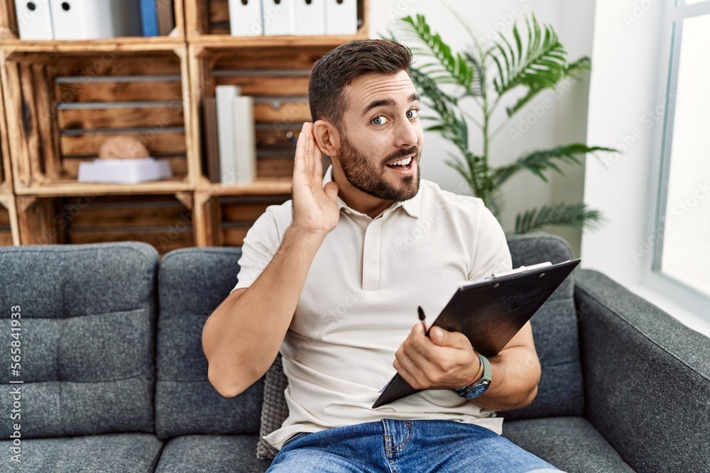 Handsome hispanic man holding clipboard working at psychology clinic smiling with hand over ear listening an hearing to rumor or gossip. deafness concept. Handsome hispanic man holding clipboard working at psychology clinic smiling with hand over ear listening an hearing to rumor or gossip. deafness concept.