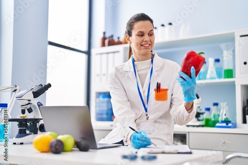 Young beautiful hispanic woman scientist holding pepper writing on document at laboratory