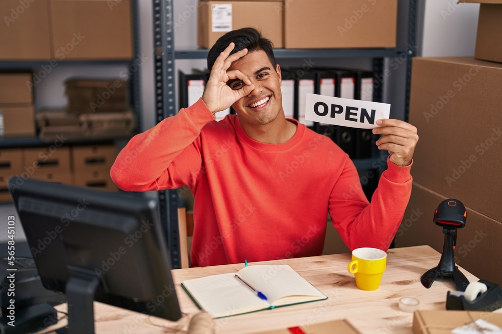 Hispanic man working at small business ecommerce holding open banner smiling happy doing ok sign with hand on eye looking through fingers