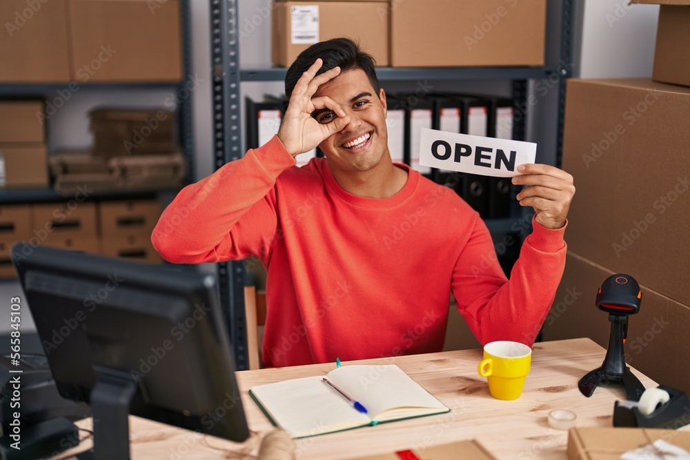 Hispanic man working at small business ecommerce holding open banner smiling happy doing ok sign with hand on eye looking through fingers Hispanic man working at small business ecommerce holding open banner smiling happy doing ok sign with hand on eye looking through fingers