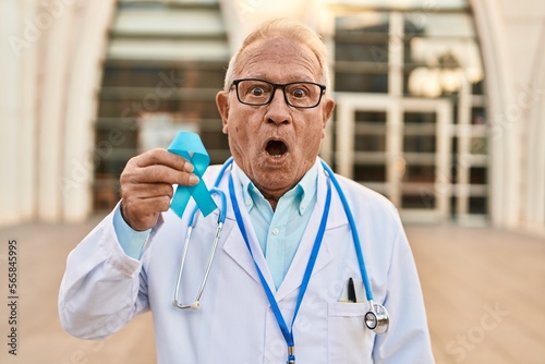 Senior doctor with grey hair holding blue ribbon scared and amazed with open mouth for surprise, disbelief face