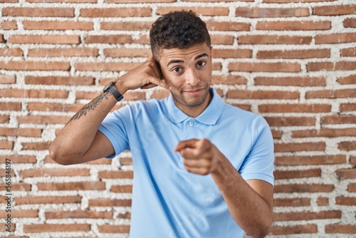 Brazilian young man standing over brick wall smiling doing talking on the telephone gesture and pointing to you. call me.