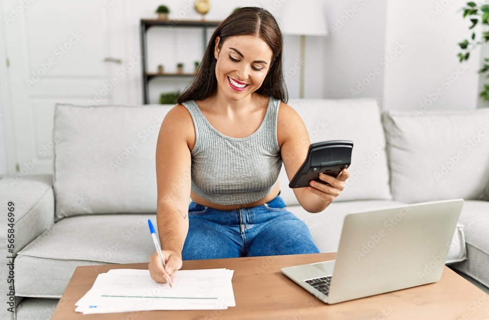 Obraz premium Young beautiful hispanic woman writing on document using calculator at home
