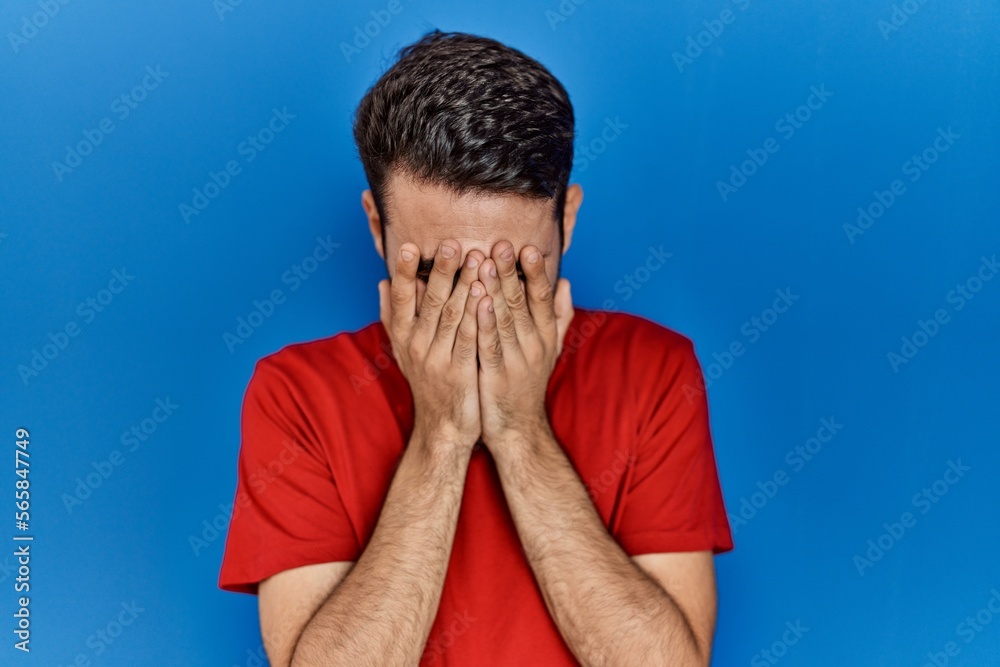 Young hispanic man with beard wearing red t shirt over blue background with sad expression covering face with hands while crying. depression concept. Young hispanic man with beard wearing red t shirt over blue background with sad expression covering face with hands while crying. depression concept.