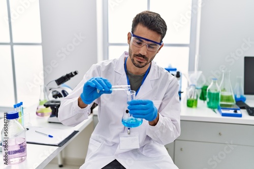 Young hispanic man wearing scientist uniform pouring liquid on test tube at laboratory