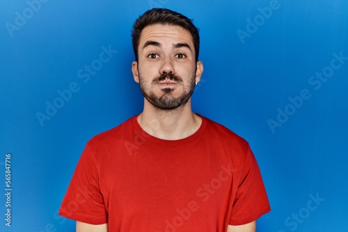 Young hispanic man with beard wearing red t shirt over blue background puffing cheeks with funny face. mouth inflated with air, crazy expression.