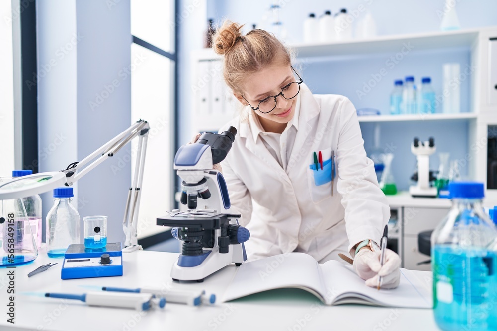 Young blonde woman scientist using microscope writing report at laboratory Young blonde woman scientist using microscope writing report at laboratory