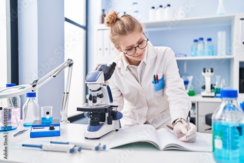 Young blonde woman scientist using microscope writing report at laboratory