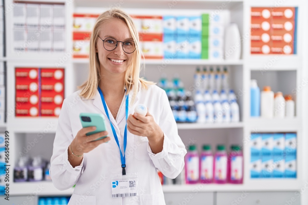 Young blonde woman pharmacist holding pills bottle using smartphone at pharmacy