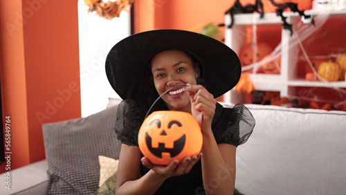 African american woman wearing witch costume holding halloween pumpkin basket at home