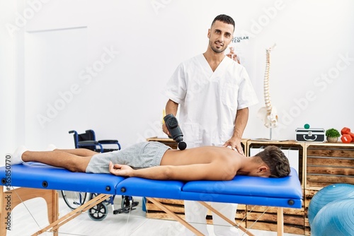 Two hispanic men physiotherapist and patient massaging back using percussion pistol at clinic