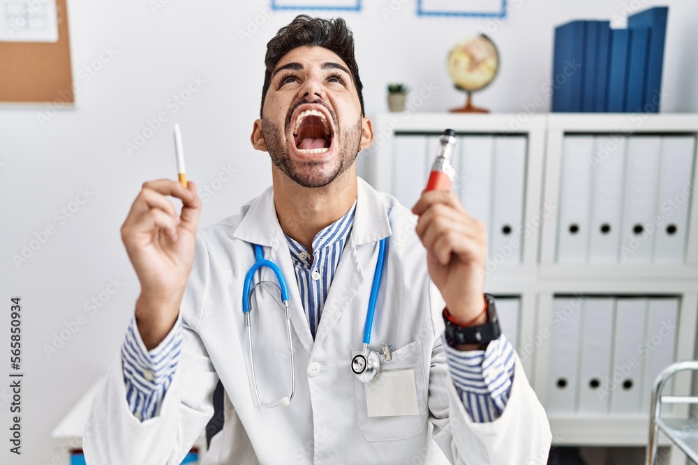 Young doctor man holding electronic cigarette at medical clinic angry and mad screaming frustrated and furious, shouting with anger looking up. Young doctor man holding electronic cigarette at medical clinic angry and mad screaming frustrated and furious, shouting with anger looking up.