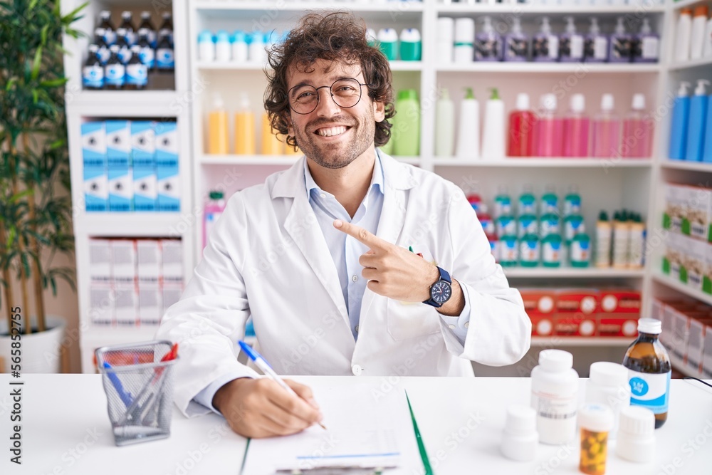 Hispanic young man working at pharmacy drugstore cheerful with a smile on face pointing with hand and finger up to the side with happy and natural expression