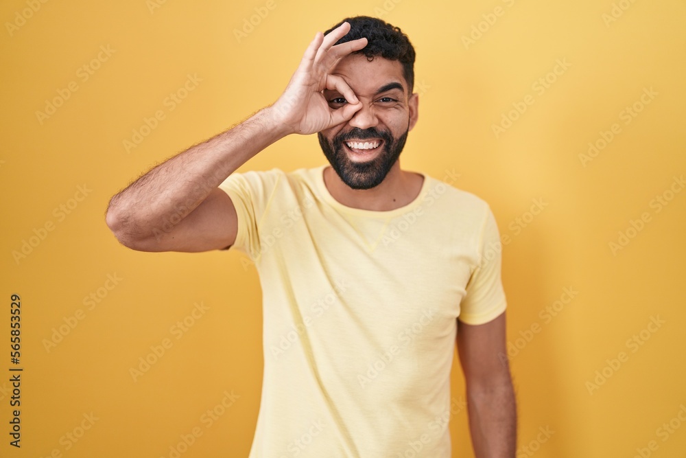 Hispanic man with beard standing over yellow background doing ok gesture with hand smiling, eye looking through fingers with happy face. Hispanic man with beard standing over yellow background doing ok gesture with hand smiling, eye looking through fingers with happy face.