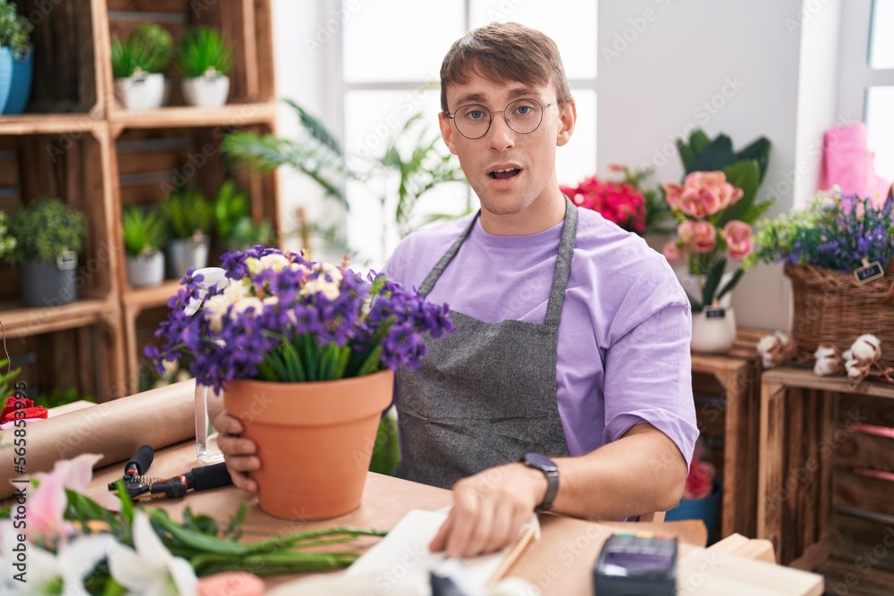 Caucasian blond man working at florist shop in shock face, looking skeptical and sarcastic, surprised with open mouth