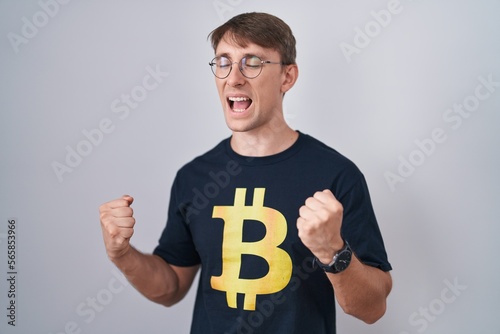 Caucasian blond man wearing bitcoin t shirt very happy and excited doing winner gesture with arms raised, smiling and screaming for success. celebration concept. © Krakenimages.com