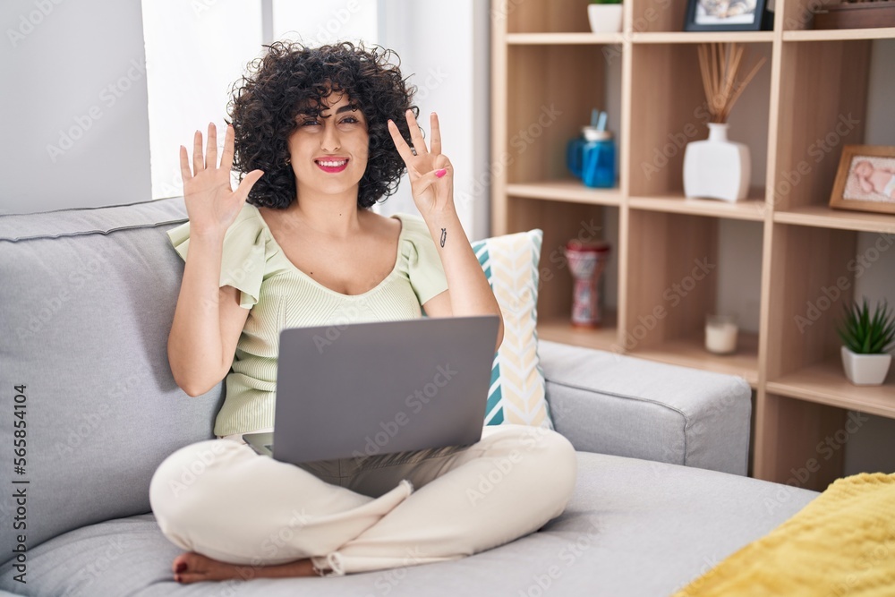 Young brunette woman with curly hair using laptop sitting on the sofa at home showing and pointing up with fingers number eight while smiling confident and happy. Young brunette woman with curly hair using laptop sitting on the sofa at home showing and pointing up with fingers number eight while smiling confident and happy.