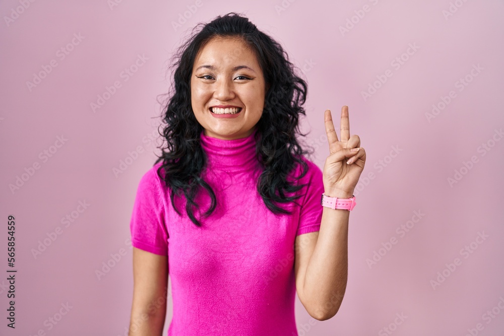 Young asian woman standing over pink background showing and pointing up with fingers number two while smiling confident and happy. Young asian woman standing over pink background showing and pointing up with fingers number two while smiling confident and happy.