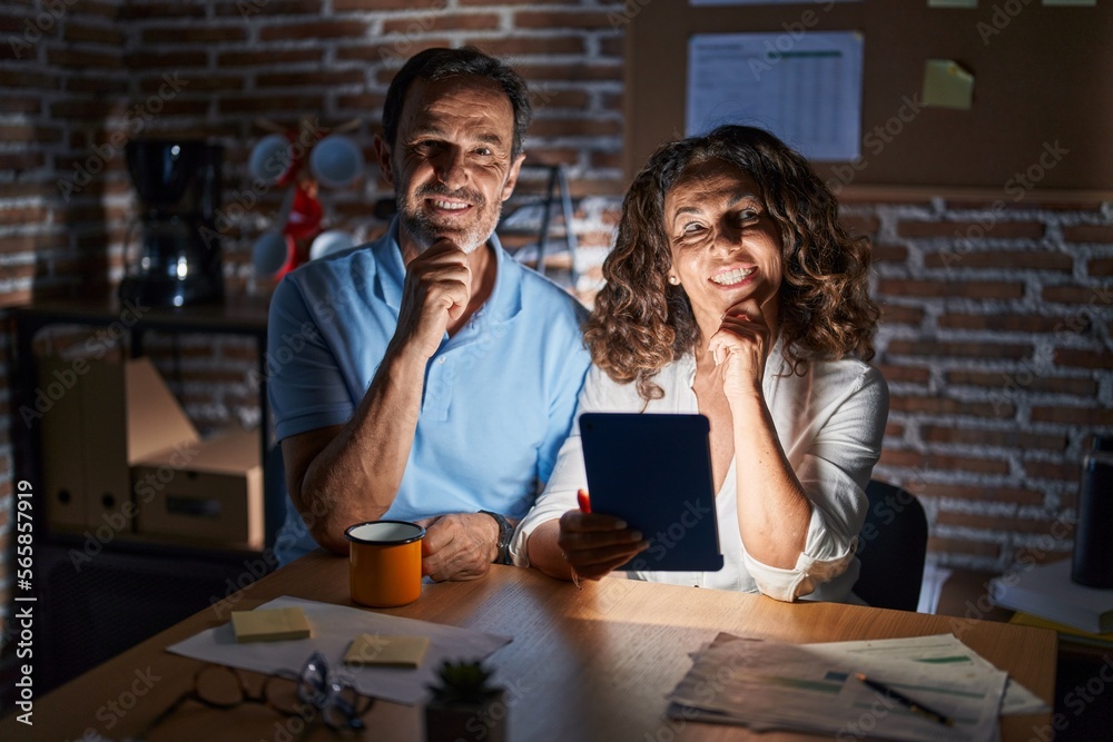 Middle age hispanic couple using touchpad sitting on the table at night with hand on chin thinking about question, pensive expression. smiling and thoughtful face. doubt concept. Middle age hispanic couple using touchpad sitting on the table at night with hand on chin thinking about question, pensive expression. smiling and thoughtful face. doubt concept.