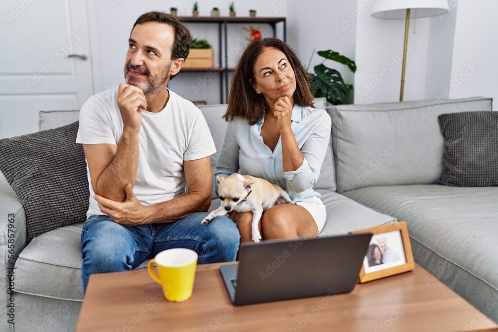 Hispanic middle age couple sitting on the sofa using computer laptop serious face thinking about question with hand on chin, thoughtful about confusing idea Hispanic middle age couple sitting on the sofa using computer laptop serious face thinking about question with hand on chin, thoughtful about confusing idea