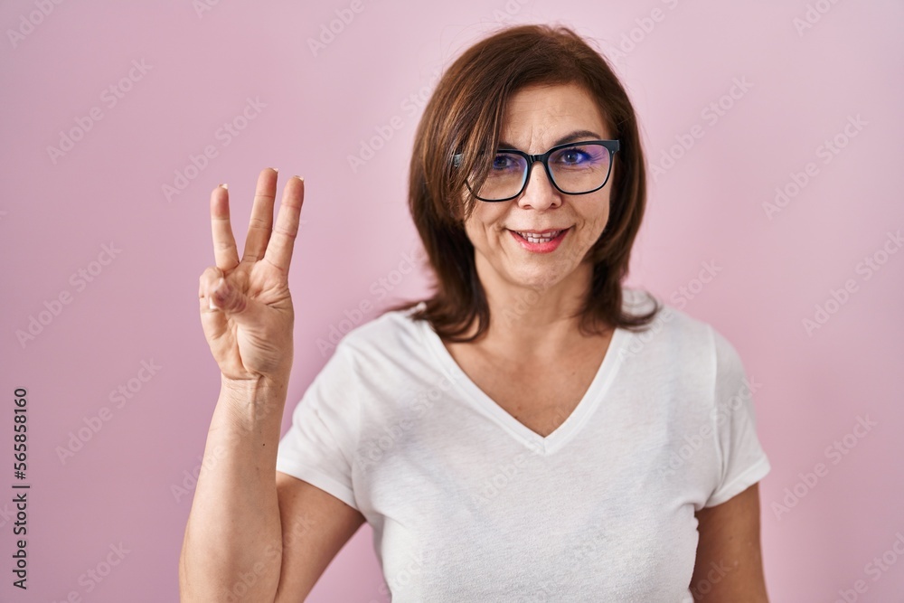 Middle age hispanic woman standing over pink background showing and pointing up with fingers number three while smiling confident and happy.