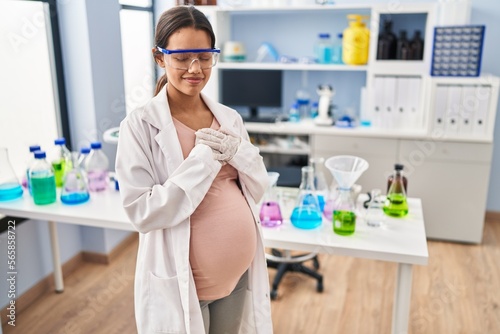 Young pregnant woman working at scientist laboratory smiling with hands on chest with closed eyes and grateful gesture on face. health concept.