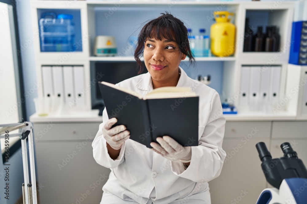 Hispanic woman working at scientist laboratory reading a book smiling looking to the side and staring away thinking. Hispanic woman working at scientist laboratory reading a book smiling looking to the side and staring away thinking.