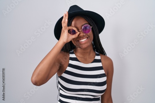 Young african american with braids wearing hat and sunglasses doing ok gesture with hand smiling, eye looking through fingers with happy face.