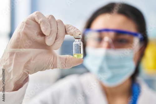 Young hispanic woman wearing scientist uniform and medical mask holding vaccine dose at laboratory