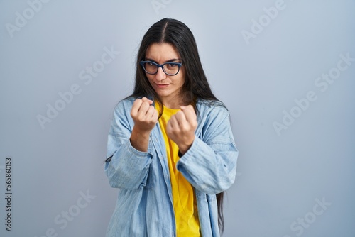 Young hispanic woman standing over blue background ready to fight with fist defense gesture, angry and upset face, afraid of problem photo