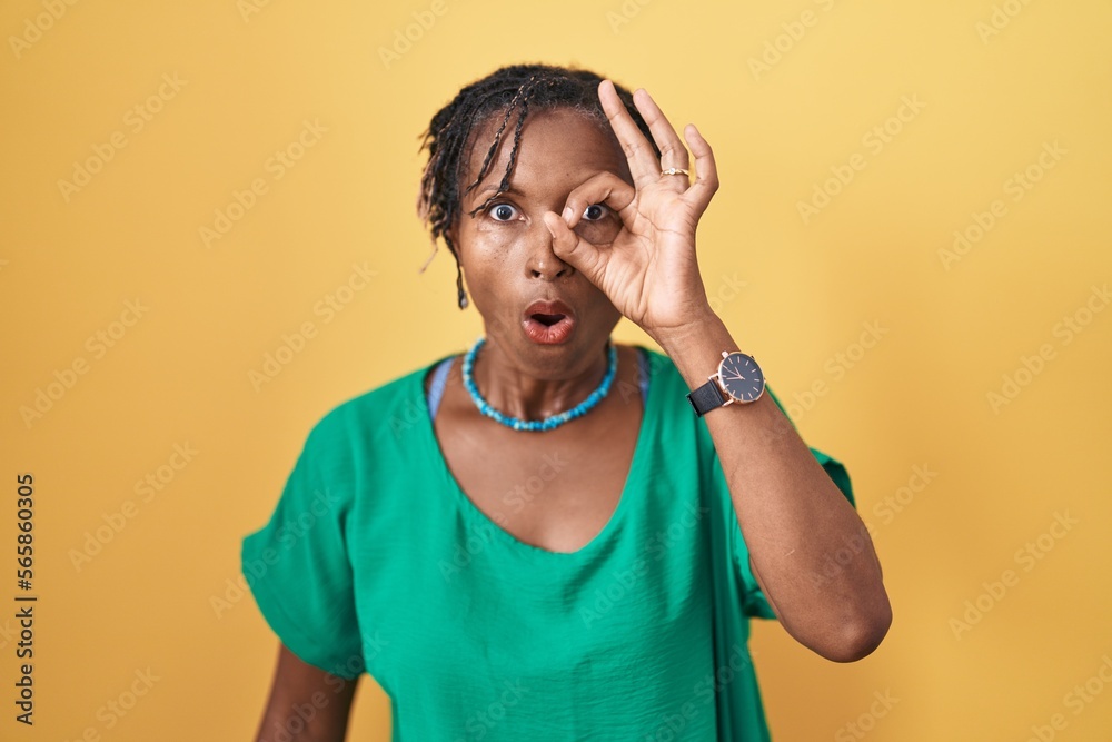 African woman with dreadlocks standing over yellow background doing ok gesture shocked with surprised face, eye looking through fingers. unbelieving expression. African woman with dreadlocks standing over yellow background doing ok gesture shocked with surprised face, eye looking through fingers. unbelieving expression.