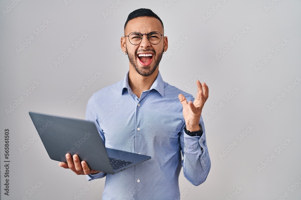 Young hispanic man working using computer laptop crazy and mad shouting and yelling with aggressive expression and arms raised. frustration concept.